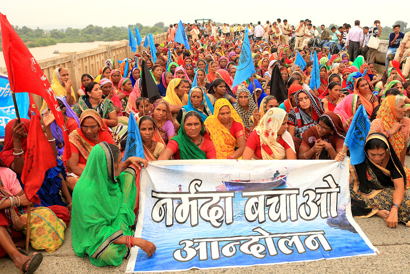 A large group of women wearing colorful traditional sarees holding banners and flags in a peaceful protest for the Narmada Bachao Andolan movement.